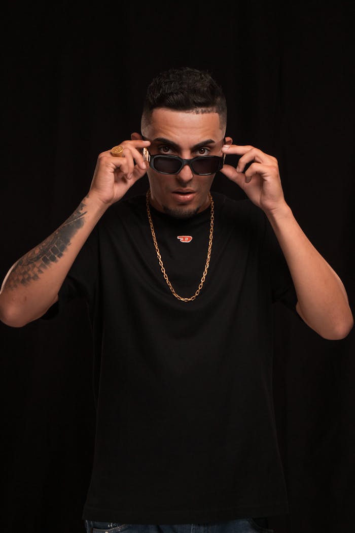 Stylish man in black t-shirt adjusting sunglasses against a dark background.